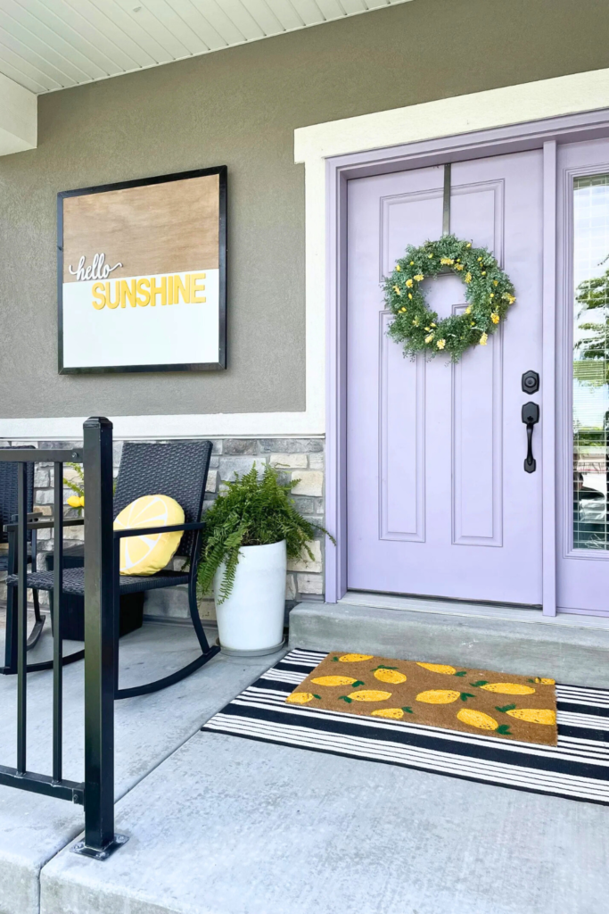 Front porch with light purple door, Hello Sunshine wood sign, black rockers, and lemon doormat