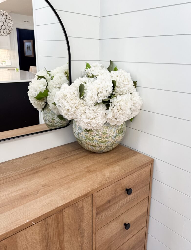 Decoupage vase with white hydrangeas sitting on a light wood entryway table with a black mirror behind it