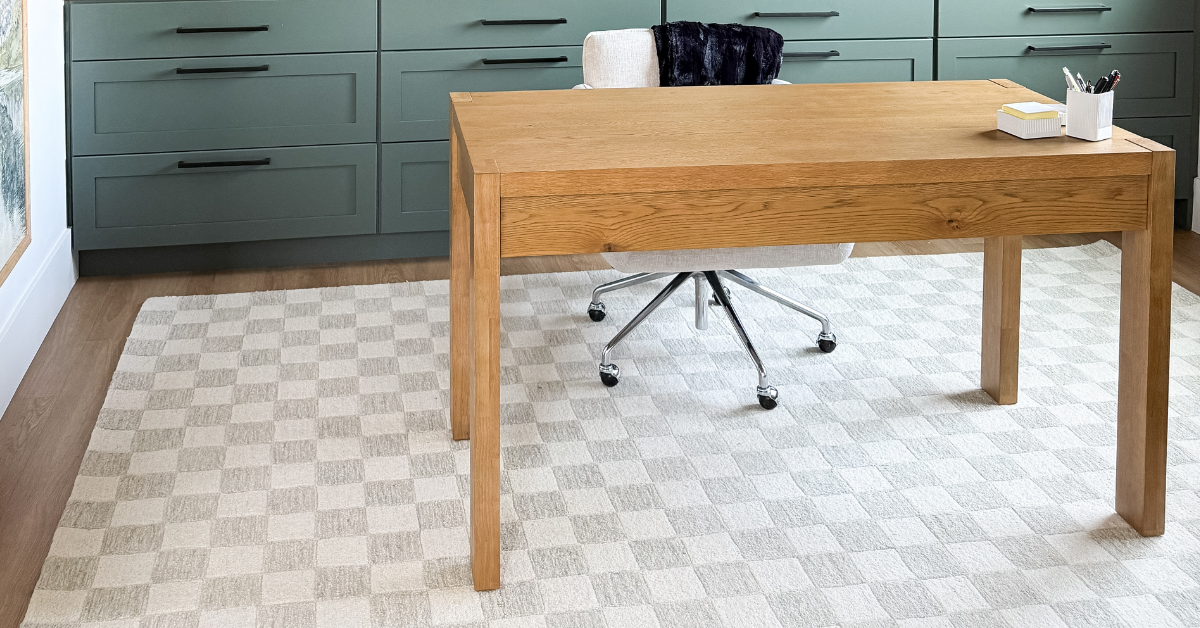 White and tan checkered rug with light wood modern desk and dark green cabinets in the background