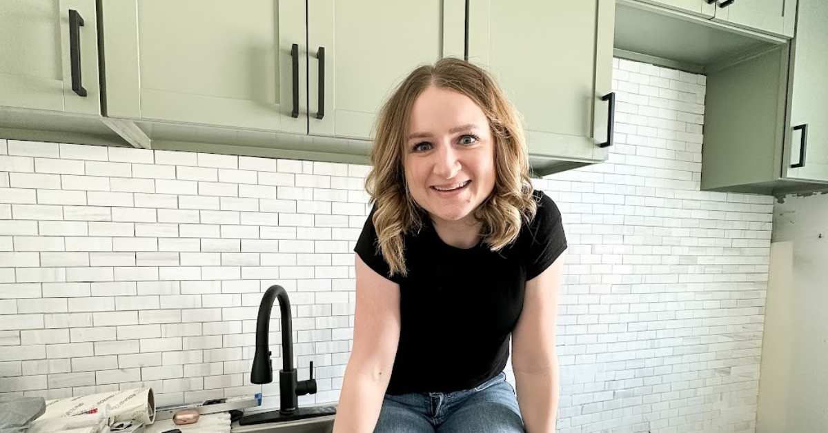 Woman sitting on the counter in front of light sage green cabinets with black pulls and a mini subway tile backsplash