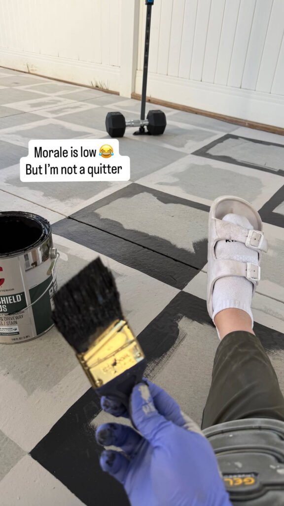 Woman sitting on the incomplete checkerboard painted patio with a paintbrush and concrete paint. Text reads: Morale is low, but I'm not a quitter.