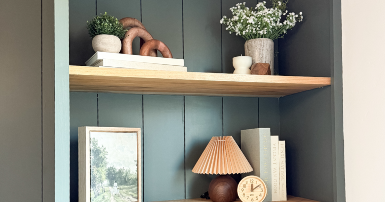 Shelf styling exmaple with art, a wood lamp, decorative books, greenery, and more on wood shelves in green built-in cabinets