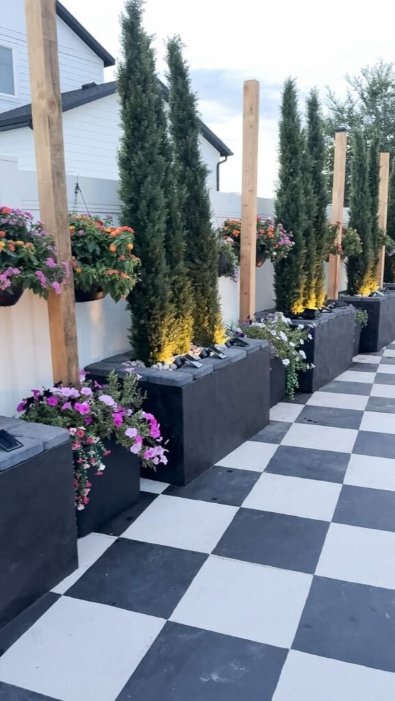 Cinder block patio planters painted black on checkerboard painted concrete. The planters are filled with tall, skinny trees, and flowers hang from the fence between the planters.