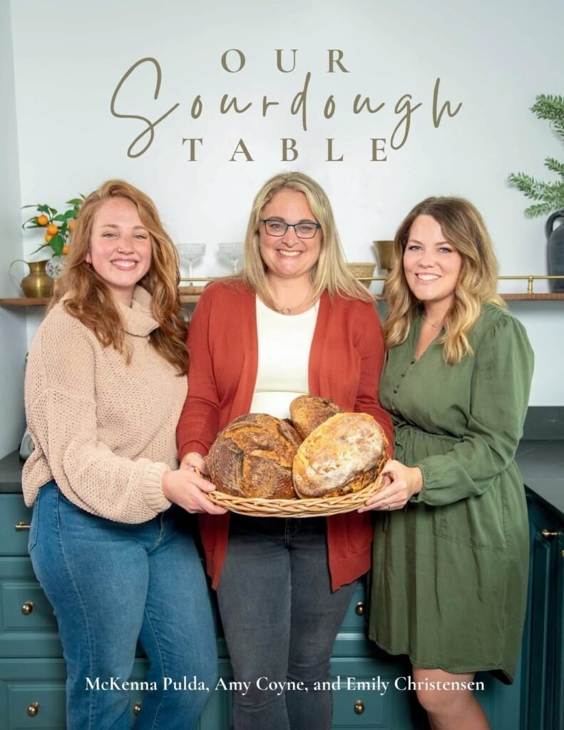Sourdough tips for beginners: get a cookbook with fun recipes! This one is Our Sourdough Table. Cover features three women holding a basket of sourdough loaves