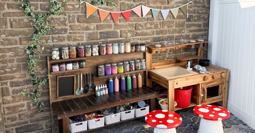 A wood mud kitchen with jars and baskets, vines on the stone fence behind it, and mini red toadstool tables
