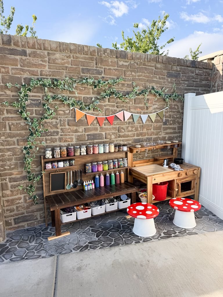 A wood mud kitchen with jars and baskets, vines on the stone fence behind it, and mini red toadstool tables