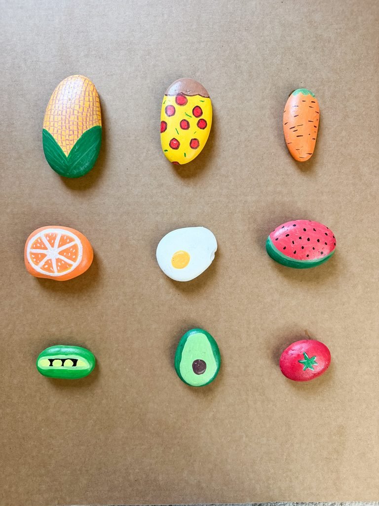 Painted rock food examples including corn, pizza, a carrot, an egg, and more