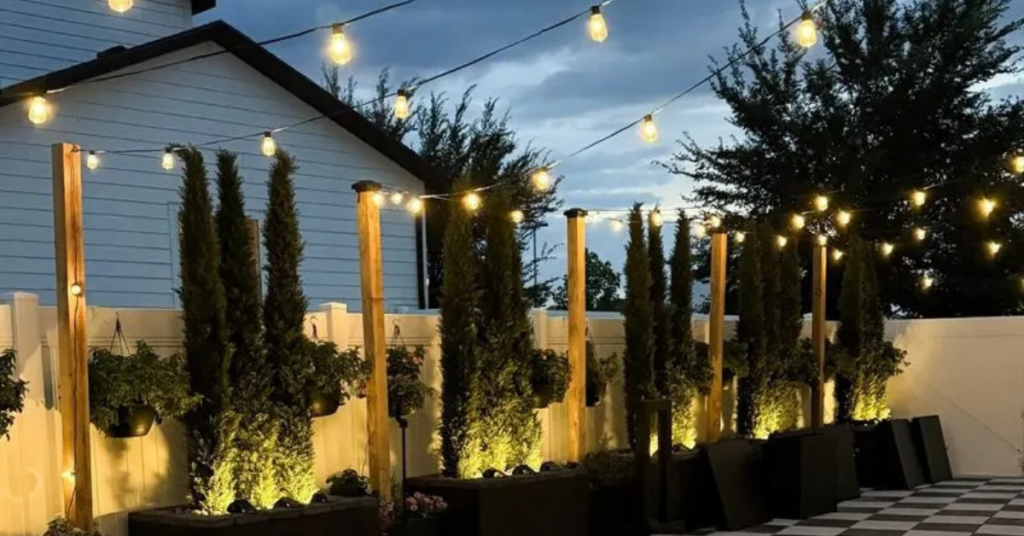 Patio transformation with wood poles and string lights hanging from them, tall privacy trees along a white vinyl fence